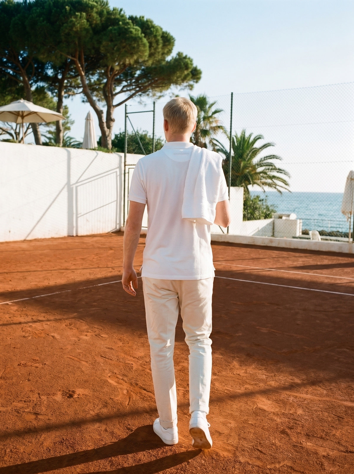A player in an Optic White polo walks off a clay court by the sea